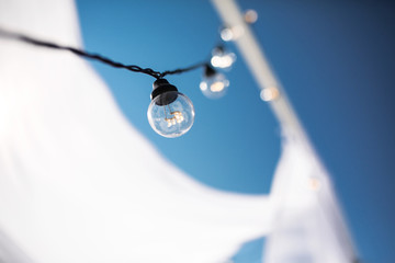 Fototapeta premium One long electric garland for lighting with white light bulbs against the background of a blue clear sky. Bokeh..Decorative wedding arch with bright bulbs
