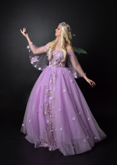  full length portrait of a blonde girl wearing a fantasy fairy inspired costume,  long purple ball gown with fairy wings,   standing pose  on a dark studio background.