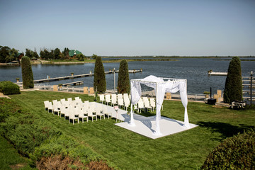Beautiful outgoing wedding set up. Jewish Hupa  on romantic wedding ceremony , wedding outdoor on the lawn water view. Wedding decor. White wooden chairs on a green lawn. White armchairs for guests