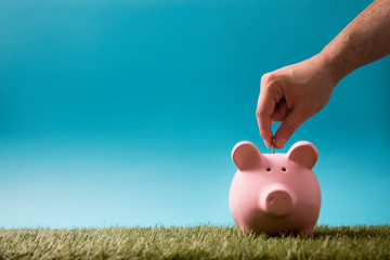 Putting coin into the piggy bank on green grass and blue sky 