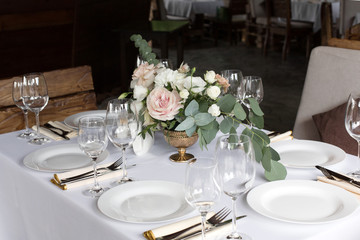Wedding table setting decorated with fresh flowers in a brass vase. Wedding floristry. Banquet table for guests outdoors with a view of green nature. Bouquet with roses, eustoma and eucalyptus leaves