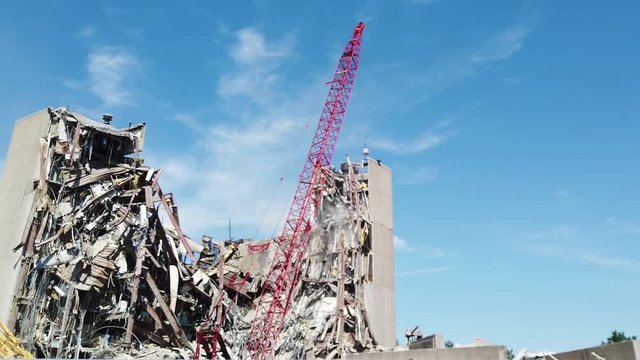Tall red crane is seen raising it's concrete wrecking ball and then lowering it onto a tower of a partially demolished building. Debris is seen to fall from the site on impact