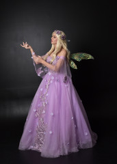  full length portrait of a blonde girl wearing a fantasy fairy inspired costume,  long purple ball gown with fairy wings,   standing pose  on a dark studio background.