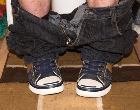 A man is sitting on the toilet in sneakers with his jeans down.A man relieves himself sitting on the toilet. - Powered by Adobe