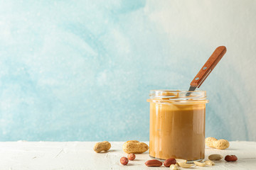 Glass jar with creamy peanut butter and spoon, and peanut on white table against light background, space for text