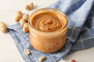 Glass jar with peanut butter, peanut and kitchen towel on white wooden background, closeup and space for text