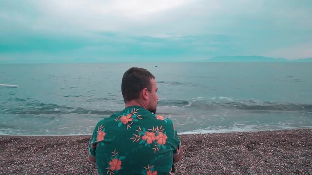 Young man sitting on the beach and looking at the sea at sunset. Handsome man  enjoys the nature and breathes fresh air in summer time. He wearing floral shirt