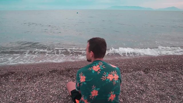 Young man sitting on the beach and  throwing pebbles into the sea. Alone man wearing floral shirt. Sunset in summer