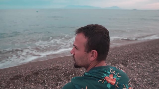 Young man sitting on the beach and  throwing pebbles into the sea. Alone man wearing floral shirt. Sunset in summer