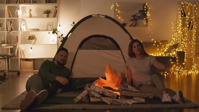 Lockdown of Caucasian man and woman sitting by artificial bonfire near tent in living room during nighttime then coming their son and daughter and sitting down near them