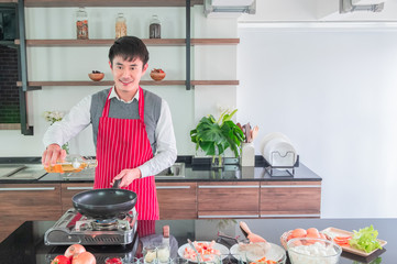 Handsome, Asian young man in Red apron, Smiling. is Pouring oil on the pan for to prepare food in...