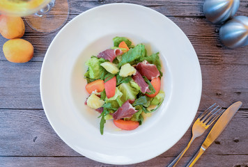 fresh summer fruit and vegetable salad with slices of prosciutto, parmesan cheese, lettuce and peach on a white plate and wooden table