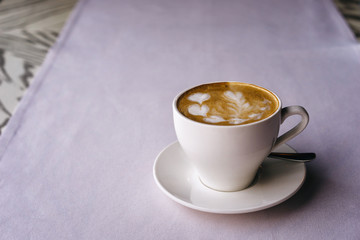 White mug on a saucer with cappuccino and beautiful heart-shaped milk foam on a purple tablecloth in a restaurant. Latte art