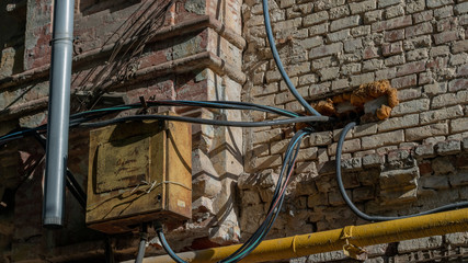 electrical metal old panel on a brick red wall. Wires and gas yellow pipe go into the house. Violation of electrical and fire safety.