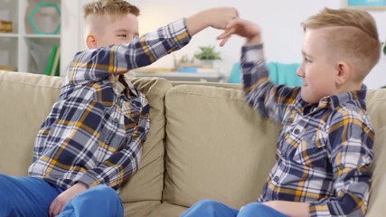 Thigh-up shot of Caucasian identical twin brothers with stylish haircuts, wearing plaid shirts, sitting on beige sofa at home, fooling around and doing fist bumps with funny hand gestures