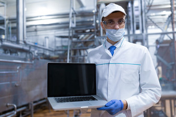 Portrait of man in a white robe and a cap standing in production department of dairy factory with laptop