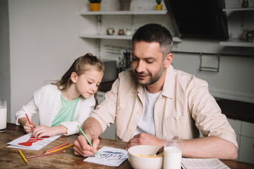 happy dad with adorable daughter drawing fathers day greeting cards while sitting at kitchen table with served breakfast