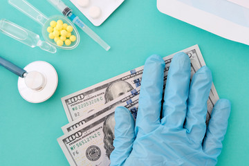A woman's hand in a blue glove on a hundred-dollar bill. Stethoscope, pills, syringe on the table. The concept of doctor's salary. Blue background.