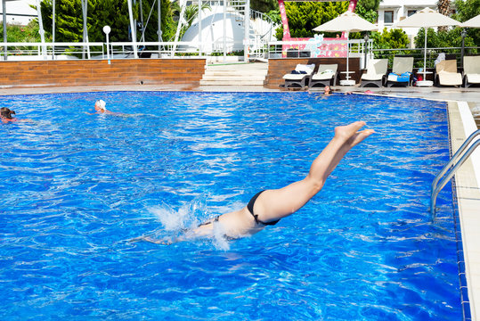 Young Brunette Jumping Off A Diving Board Into An Outdoor Swimming Pool. Woman In Black Bikini Doing Caught In Mid Air Doing A Cannon Ball Into A Swimming Pool In The Backyard.