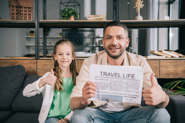 smiling father and daughter showing thumbs up while sitting on sofa with travel life newspaper