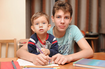 Obraz premium Two brothers of different ages sit at the table at home and draw, the older brother teaches to draw a younger brother