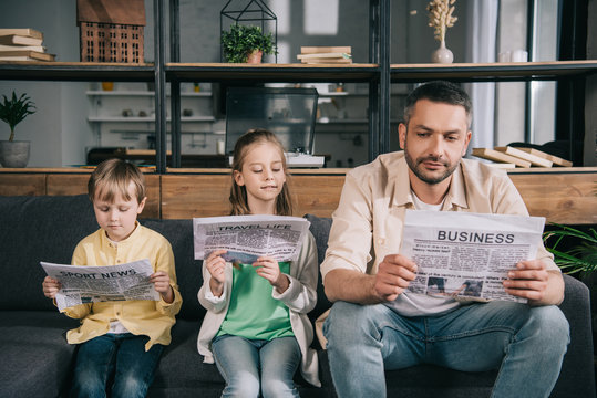 Father With Adorable Kids Reading Newspapers While Sitting On Sofa At Home