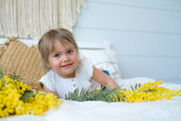 A beautiful little girl is lying on the bed next to a bouquet of Mimosa. The baby has blonde hair and blue eyes. White bedroom, cozy house. Spring holiday concept