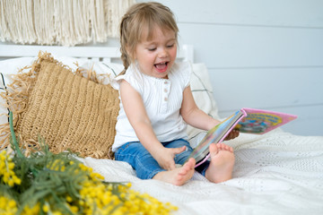 Beautiful little girl sitting on the bed and reading a book. The baby has blonde hair and blue eyes. White bedroom, cozy house. Teaching children to read.