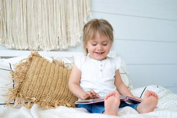 Beautiful little girl sitting on the bed and reading a book. The baby has blonde hair and blue eyes. White bedroom, cozy house. Teaching children to read.