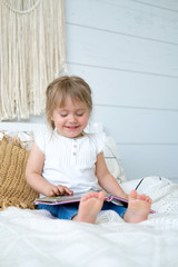Beautiful little girl sitting on the bed and reading a book. The baby has blonde hair and blue eyes. White bedroom, cozy house. Teaching children to read.