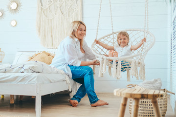 Beautiful little girl swinging in a hanging chair in the bedroom. The child's mother is sitting next to him on the bed. Macrame wicker chair, knitted blanket, straw pillow. Cozy home, happy family
