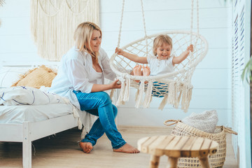 Beautiful little girl swinging in a hanging chair in the bedroom. The child's mother is sitting next to him on the bed. Macrame wicker chair, knitted blanket, straw pillow. Cozy home, happy family