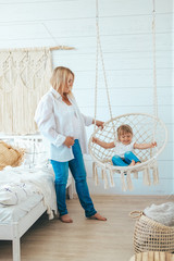 Beautiful little girl swinging in a hanging chair in the bedroom with her mom. A woman is pregnant with her second child. Macrame wicker chair, knitted blanket, straw pillow. Cozy home, happy family