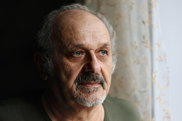 Closeup portrait of sad old man indoor: senior man with grey beard and moustache standing near window. Loneliness, aged people problems, fear of future, depression, uncertainity in the future concept