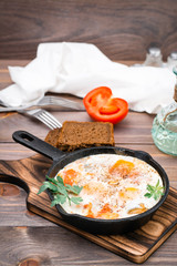 Homemade breakfast of shakshuka fried eggs with tomatoes and herbs in a pan on a cutting board on a wooden table
