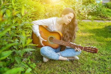 Young hipster woman sitting in grass and playing guitar on park or garden background. Teen girl...