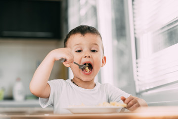 a child in the kitchen during the day eating pasta in a spiral in a white t-shirt