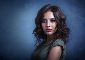 Naklejka premium Beautiful makeup brunette model with curly hair looking sexy on gray-blue dark background. Close-up of a woman portrait.