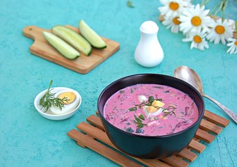 Cold beet soup with green onions, cucumber and dill on kefir or buttermilk in a black bowl on a turquoise background. Served with boiled egg. Lithuanian cuisine. Healthy food, diet.
