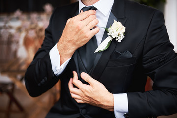 Elegnat groom in black stylish suit putting on a tie in the morning