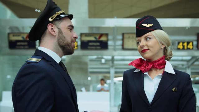 In airport stewardess with blond hair smiling affably, talking with pilot. European woman dressed in dark blue uniform, scarlet red scarf tied on her white shirt. Cinematic shot on RED camera.