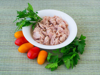Canned tuna fillet in white porcelain bowl, parsley and some cherry tomatoes on a green table mat made of natural plant fibers. Seafood, healthy eating.