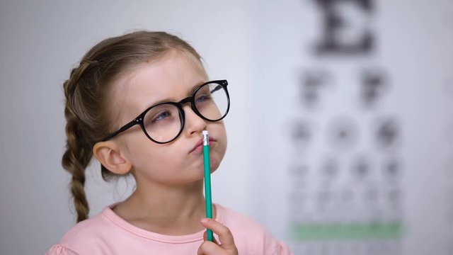 Pensive smart girl wearing glasses, testing vision with eye chart in clinic