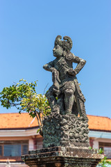 Fototapeta na wymiar Klungkung, Bali, Indonesia - February 26, 2019: Closeup of Stone statue of defiant King covered in black mold against blue sky. Some green foliage and red roof.