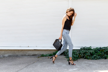 Young stylish woman wearing black cami top, blue cropped denim jeans, black high heel sandals and holding bag walking in the city street. Trendy casual outfit for summer or spring. Street fashion.
