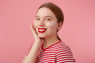 Young smiling red-haired girl with red lips and with patches under the eyes, wears in a red striped T-shirt, touches the cheek, stands over pink background and enjoying free time for skin care.