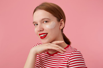 Nice smiling red-haired girl wears in a red striped T-shirt, with red lips and with patches under the eyes, touches the cheen, stands over pink background and enjoying free time for skin care. © timtimphoto