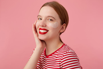 Young smiling red-haired lady with red lips wears in a red striped T-shirt, with patches under the eyes,touches the cheek,enjoying free time for skin care and stands over pink background.