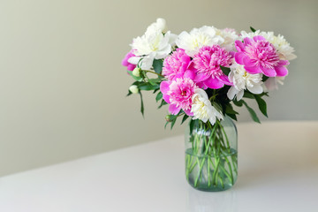 Bouquet of fresh big pink, white and cream peonies in simple glass jar on glance table indoor. Vase with beautiful tender spring flowers on glass table