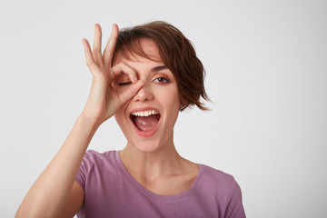Close up of young attractive happy short-haired woman in blank t-shirt, looking through okay gesture and broadly smiling, standing over white wall .Positive emotion concept.